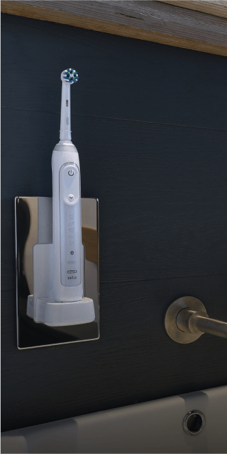 Proofvision - A white electric toothbrush with a round brush head is mounted on a chrome-colored wall holder. The holder is attached to a dark-colored wall, and a silver towel ring is visible nearby. The toothbrush is charging on the holder.