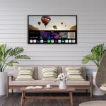 Proofvision - A flat-screen TV is mounted on a white shiplap wall, displaying a colorful hot air balloon scene and various app icons on the home screen. Below the TV, there are green potted plants, a beige sofa is visible.
