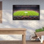 Proofvision - A TV mounted on a white brick wall displays a soccer stadium with fans and players. Below, there is a wooden table and to the right, a wheelbarrow with plants and gardening tools. A closed wooden shutter is on the left side of the wall.