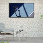 Proofvision - A modern living room with white brick walls features a light blue cushioned chair and matching ottoman next to a small white table. Above the furniture, a large framed photo of a tennis player in action is mounted on the wall. Plants adorn both sides of the space.