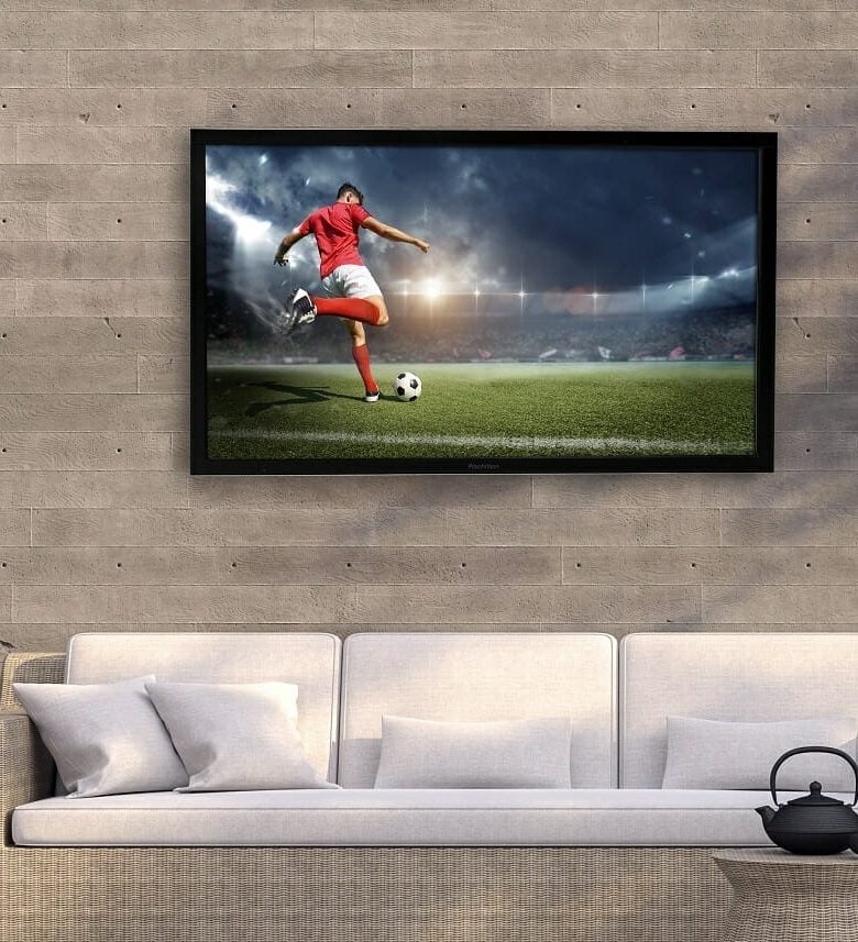 Proofvision - A flat-screen TV mounted on a wall above a light grey sofa. The TV displays an image of a soccer player in a red jersey and white shorts, kicking a soccer ball on a grassy field under stadium lights. A black teapot sits on a small round table in front of the sofa.