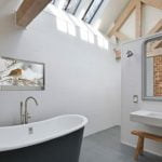 Proofvision - A minimalist bathroom features a freestanding black and white bathtub, a wall-mounted sink, a large mirror with a gray frame reflecting a brick wall, and a wooden bench. A skylight and a bird picture on the wall add light and a touch of nature to the space.