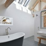 Proofvision - Modern bathroom with a freestanding oval bathtub, minimalist sink with wooden bench, large mirror, and skylights. Walls feature exposed wooden beams and brick details, and there is a framed picture of a bird on a branch above the bathtub.
