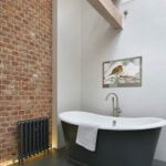 Proofvision - A modern bathroom featuring a freestanding black bathtub with a white interior, a towel hanging over the edge, and a chrome faucet. The room has an exposed brick wall, a black radiator, a large painting of a bird, and a wooden ladder leaning against the white wall.