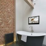 Proofvision - A modern bathroom with a free-standing black bathtub under a tall white wall. A white towel hangs over the bathtub's edge. A brick accent wall features a radiator beneath. A large ladder leans against the white wall, near a framed picture of a bird.