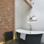 Proofvision - A modern bathroom featuring a standalone black bathtub with a towel draped over the edge. One wall is exposed red brick, and the other is white with a bird photo hanging. A tall metallic ladder leans against the white wall, and a black radiator is below the brick wall.