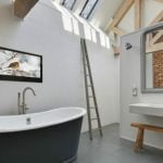 Proofvision - A modern bathroom with a freestanding bathtub, wall-mounted sink, large mirror, and exposed wooden beams. A metal ladder leans against the wall, and a picture of a bird in winter hangs on the wall. The ceiling has skylights, and one wall has exposed bricks.