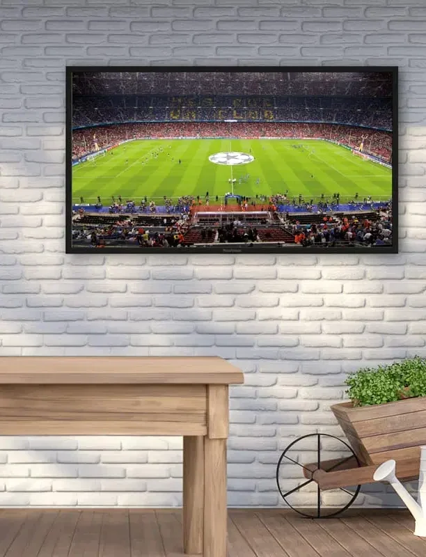 Proofvision - A television mounted on a white brick wall displays a soccer match in a stadium with a central logo on the pitch. Below the TV is a wooden table and a decorative cart with green plants.