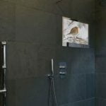 Proofvision - A modern bathroom with dark slate tiles, featuring a wall-mounted television screen displaying a bird image. There is a heated towel rail on the left, a rainfall showerhead on the right, and a "SMART TV" label in the bottom right corner.