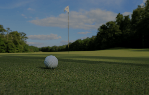 Proofvision - A close-up of a golf ball sitting on a manicured green with a golf hole and flagstick visible in the background. The scene is set on a sunny day, with trees and a blue sky with clouds in the distance.