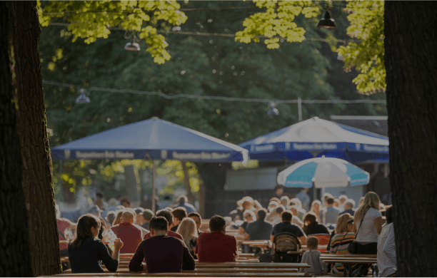 Proofvision - Outdoor scene at a bustling beer garden with people seated at wooden tables under the shade of large trees. Blue and white umbrellas provide additional shade, and warm sunlight filters through the branches, creating a relaxed and inviting atmosphere.