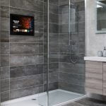 Proofvision - A modern bathroom features a glass-enclosed shower with dark gray tiles, a mounted TV screen, and a sleek hand shower. The vanity area includes a light wood cabinet with a white sink, a large mirror, and soap dispensers. Open shelves hold decorative items.