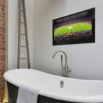 Proofvision - A modern bathroom featuring a white freestanding tub with a towel draped over the side, a sleek faucet, and a ladder-style shelf. A brick wall is on the left side, and a large flat-screen TV showing a soccer match is mounted on the adjacent wall.