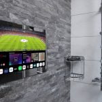 Proofvision - A flat-screen TV is mounted on a gray tiled bathroom wall above a stainless steel shower caddy and near a showerhead. The TV screen displays a soccer match and various app icons at the bottom. The surrounding tiles are a mix of dark and light gray shades.
