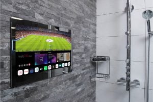 Proofvision - A flat-screen TV is mounted on a gray tiled bathroom wall above a stainless steel shower caddy and near a showerhead. The TV screen displays a soccer match and various app icons at the bottom. The surrounding tiles are a mix of dark and light gray shades.