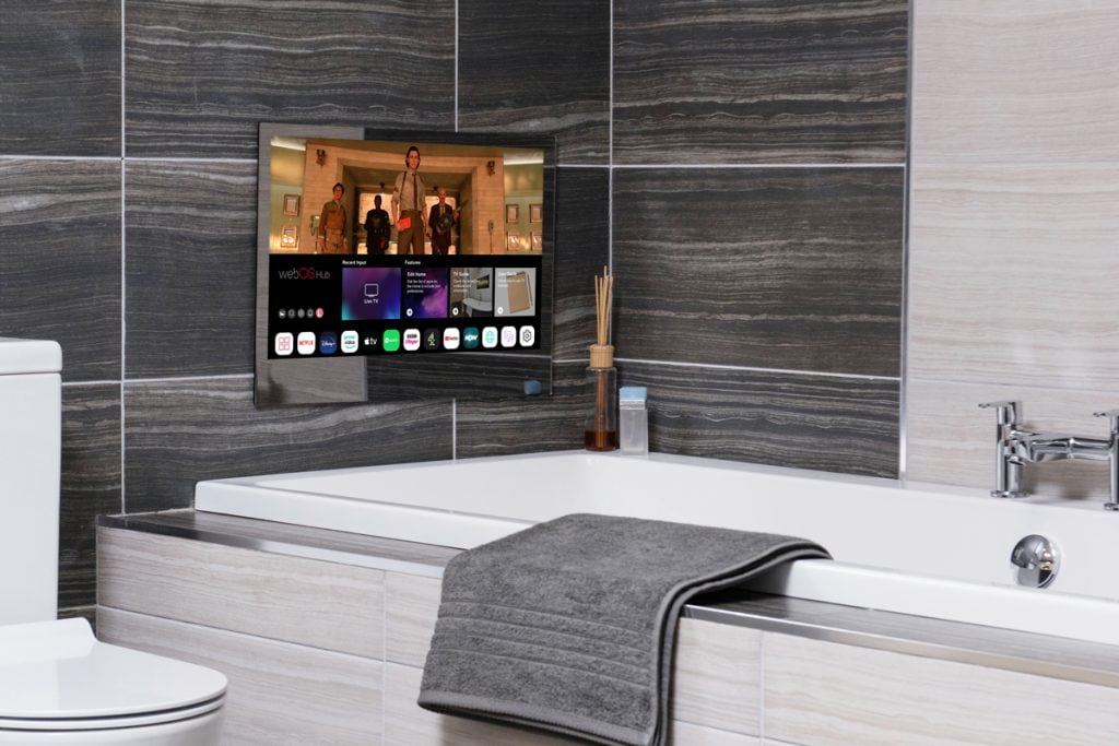 Proofvision - A modern bathroom featuring a TV integrated into the tiled wall above a bathtub. The TV screen displays various apps and a movie. The bathtub has a gray towel draped over its edge. The backdrop includes sleek gray and beige tiles, and the toilet is visible to the left.