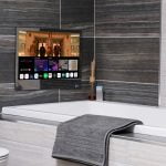 Proofvision - A modern bathroom featuring a TV integrated into the tiled wall above a bathtub. The TV screen displays various apps and a movie. The bathtub has a gray towel draped over its edge. The backdrop includes sleek gray and beige tiles, and the toilet is visible to the left.