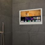 Proofvision - A modern bathroom with gray tiled walls features a flat-screen TV mounted on one wall. The TV displays a nature scene with a fox, as well as a menu of various streaming and entertainment options below. A silver showerhead and faucet are mounted on the adjacent wall.